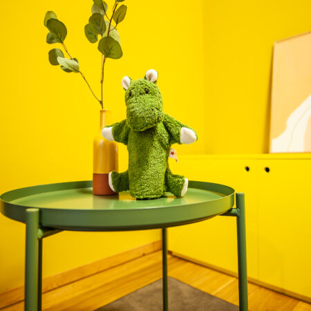 Grünes Plüschnilpferd und Vase mit Pflanze auf grünem Tisch vor gelber Wand