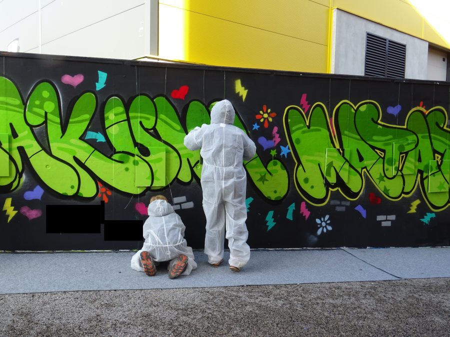 Graffiti-Sprayer in weißen Schutzanzügen vor bunter Wand mit Schriftzug